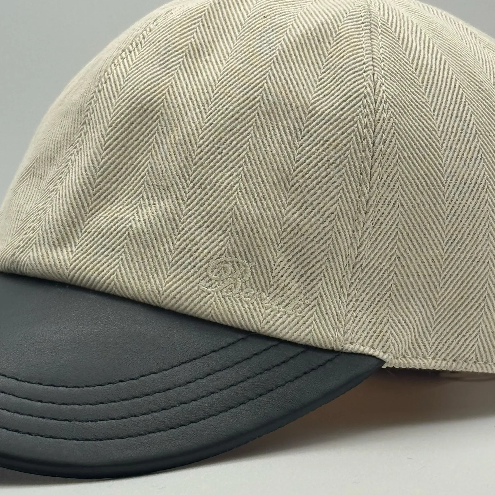 Close-up of the black leather brim meeting the herringbone linen crown, with a small tonal logo at the seam.